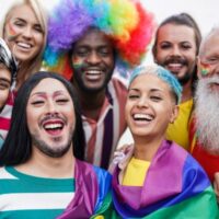 7 people of different ages - older and younger - and different races and genders/gender expressions with Pride flags, rainbow wigs and body art.