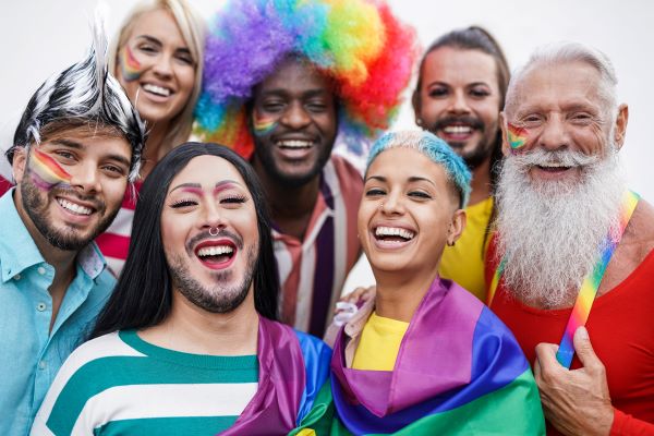 7 people of different ages - older and younger - and different races and genders/gender expressions with  Pride flags, rainbow wigs and body art.